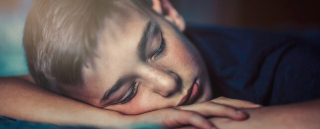 Boy with light brown hair taking nap on forearm at desk.