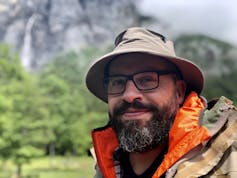 Man in hat, glasses and bear smiles at camera, forest in background