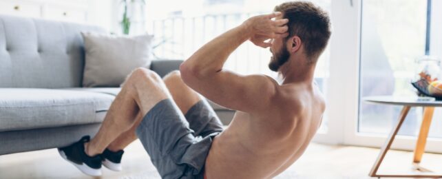 A topless masculine person doing crunches in a lounge room